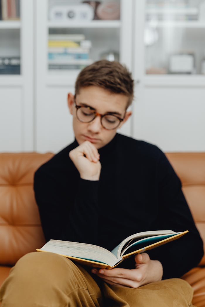 Teenager wearing glasses reading a book on a couch, exuding intelligence.
